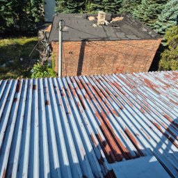 FULMAL Piotr Górka - Widok z góry na pordzewiały, częściowo pomalowany dach z blachy falistej, z budynkiem z czerwonej cegły w tle i zielenią drzew. Widoczne ślady rdzy i niedokładnego malowania.