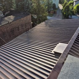 FULMAL Piotr Górka - Nowy, brązowy dach z blachy trapezowej, widok z góry na tle zieleni i zabudowań. Widoczne detale łączeń i mocowań. Słoneczny dzień.