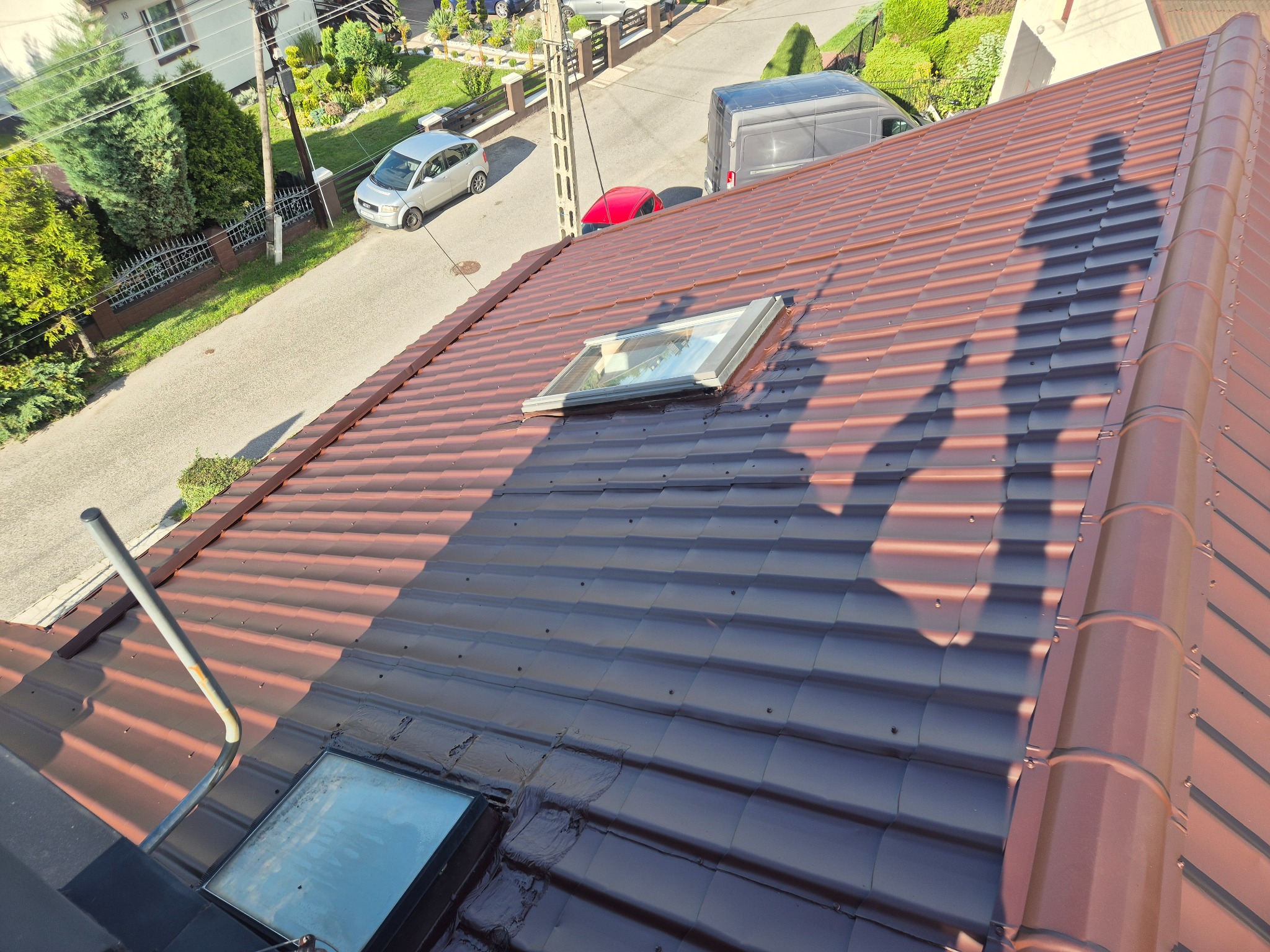 Częściowo pomalowany dach z dachówki ceramicznej w odcieniach brązu i grafitu, z widocznymi oknami dachowymi i fragmentem ulicy w tle.