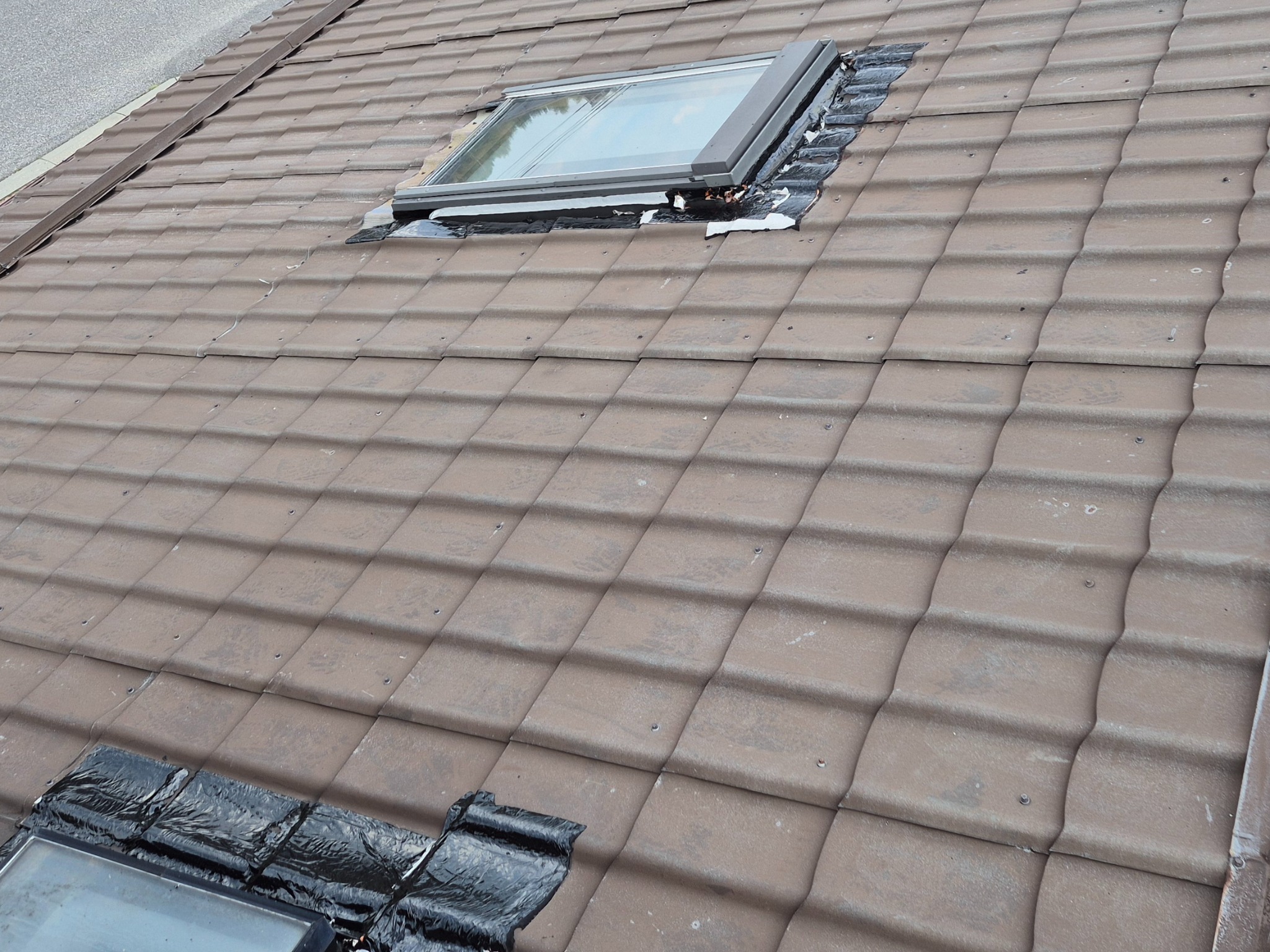 Brązowa dachówka ceramiczna z oknem dachowym w trakcie uszczelniania czarną folią. Widoczne ślady po montażu i zabrudzenia na powierzchni dachu.