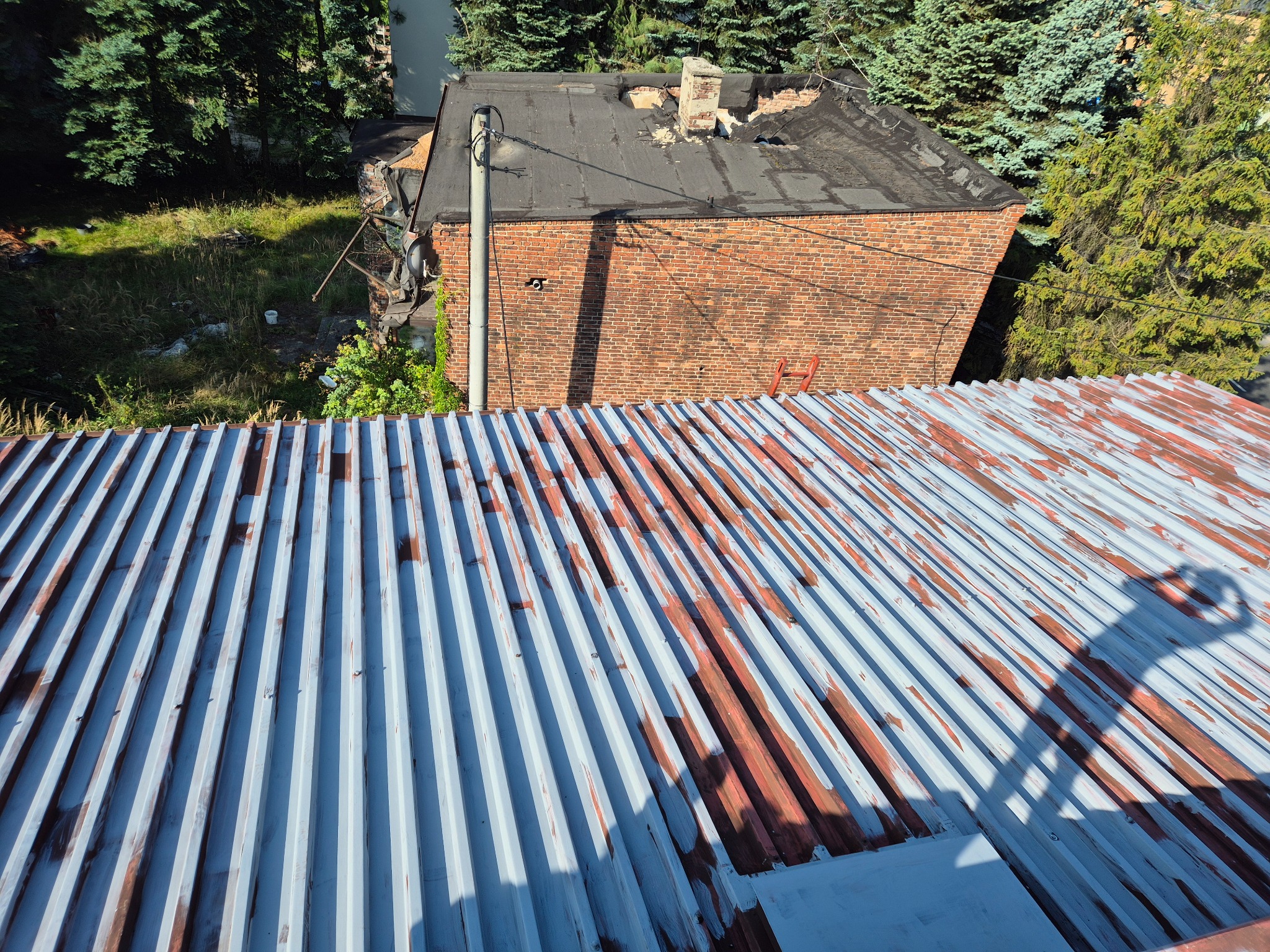 Widok z góry na pordzewiały, częściowo pomalowany dach z blachy falistej, z budynkiem z czerwonej cegły w tle i zielenią drzew. Widoczne ślady rdzy i niedokładnego malowania.