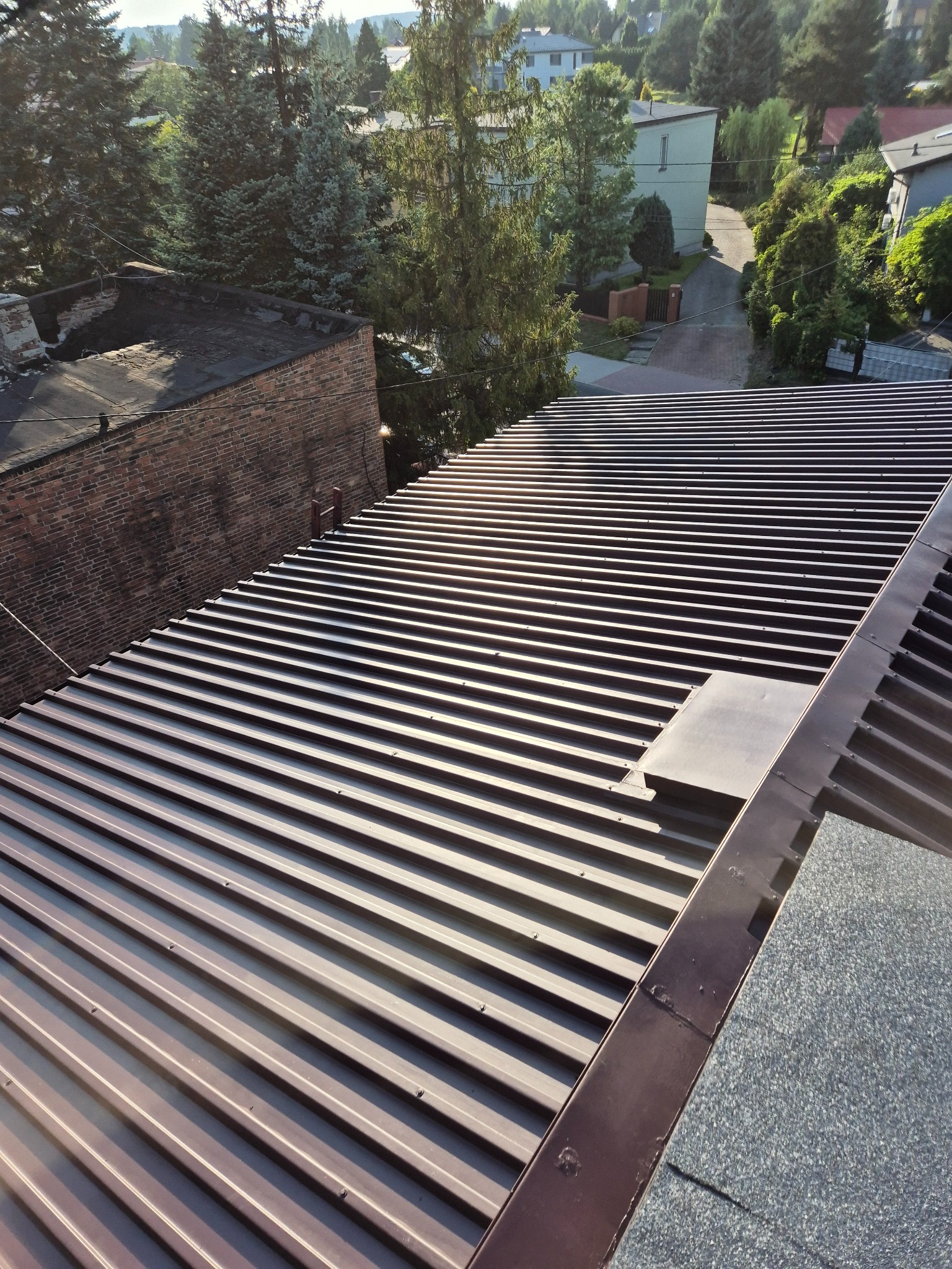 Nowy, brązowy dach z blachy trapezowej, widok z góry na tle zieleni i zabudowań. Widoczne detale łączeń i mocowań. Słoneczny dzień.