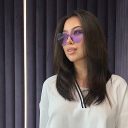 IBBEAUTY Sp.z o.o. - Portret kobiety w okularach w kształcie serc na tle zasłon. Uczesanie z przedziałkiem, delikatny makijaż, jasna bluzka. Ujęcie z boku, oświetlenie naturalne.