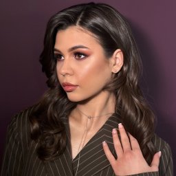 IBBEAUTY Sp.z o.o. - Portret kobiety z profesjonalnym makijażem oczu i ust, brązowe włosy ułożone w fale, ręka na ramieniu w brązowym żakiecie w prążki, fioletowe tło.