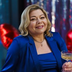 IBBEAUTY Sp.z o.o. - Uśmiechnięta blondynka w niebieskim żakiecie, z kieliszkiem szampana na tle błyszczących dekoracji i czerwonych balonów w kształcie serc.