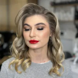 Portret kobiety z profesjonalnym makijażem oczu i czerwonymi ustami, oraz blond włosami ułożonymi w fale w stylu retro. Widoczne srebrne kolczyki.