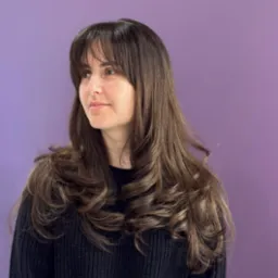 Portret kobiety z długimi, brązowymi, kręconymi włosami z grzywką na tle fioletowej ściany. Włosy po stylizacji fryzjerskiej, ułożone w fale.