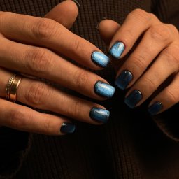 IBBEAUTY Sp.z o.o. - Dłonie z kwadratowymi paznokciami, pomalowanymi lakierem w odcieniach granatu i błękitu z efektem 'kocie oko', na tle brązowego swetra.