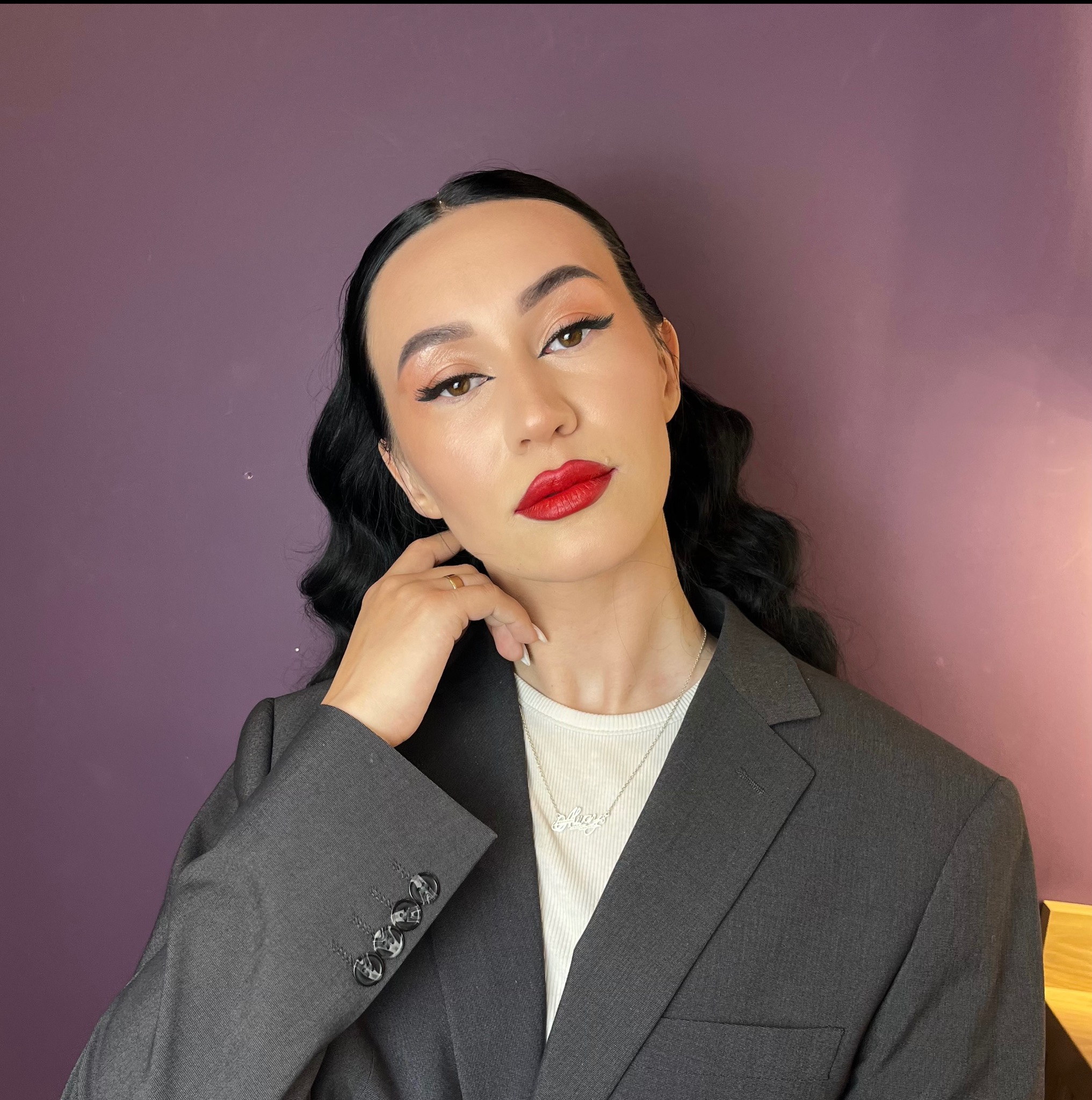 Kobieta z wyrazistym makijażem oczu i ust w odcieniu czerwieni, pozująca w szarym żakiecie na tle fioletowej ściany. Uwagę przykuwa precyzyjny eyeliner i podkreślone brwi.