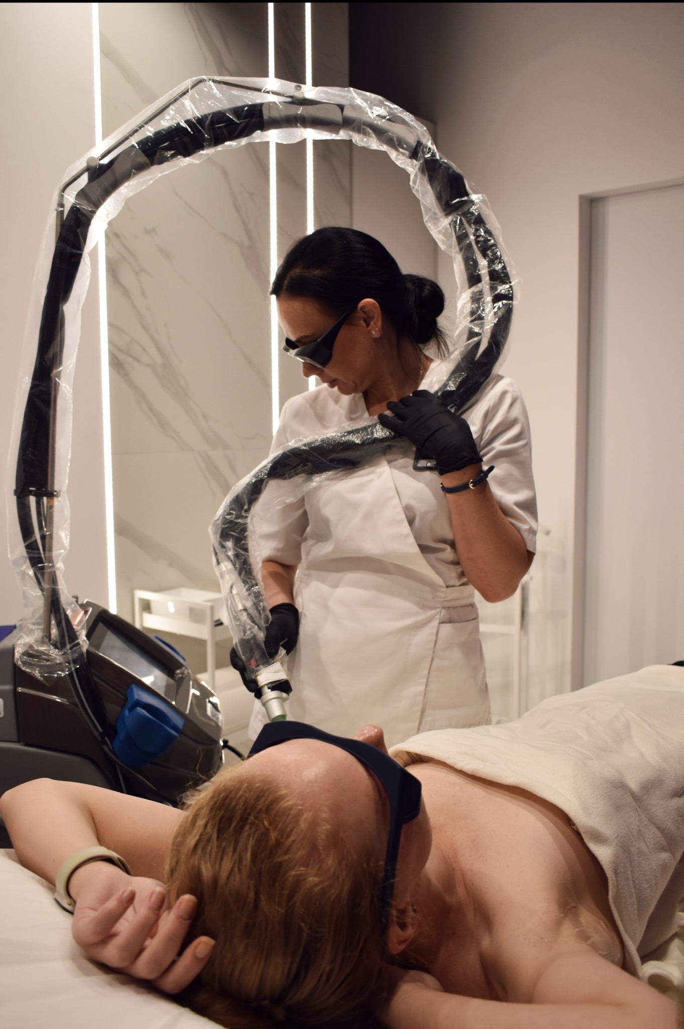 Klientka z ochroną na oczach podczas zabiegu depilacji laserowej. Kosmetolog w okularach ochronnych obsługuje urządzenie. Nowoczesny salon.