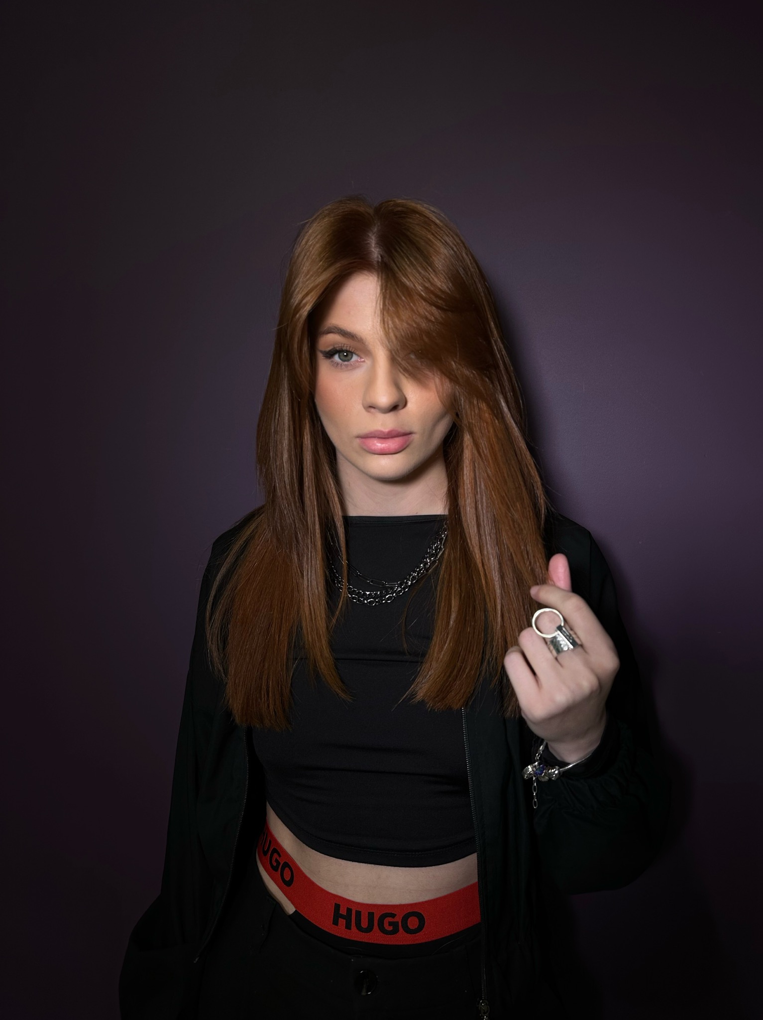 Portret kobiety z rudymi włosami, w czarnym ubraniu, na fioletowym tle. Włosy opadają na twarz, widoczny makijaż oczu. Uwagę zwraca duży pierścień na palcu.