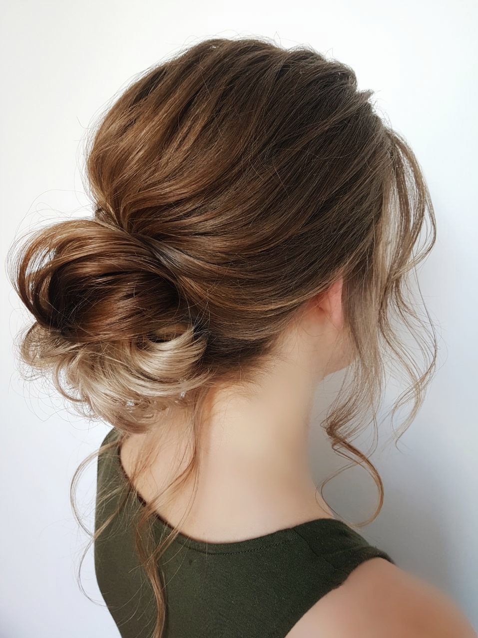 Upięcie włosów w kok z luźnymi pasmami, widok z tyłu, brązowe włosy z blond refleksami, gładka skóra, zielona bluzka, białe tło.