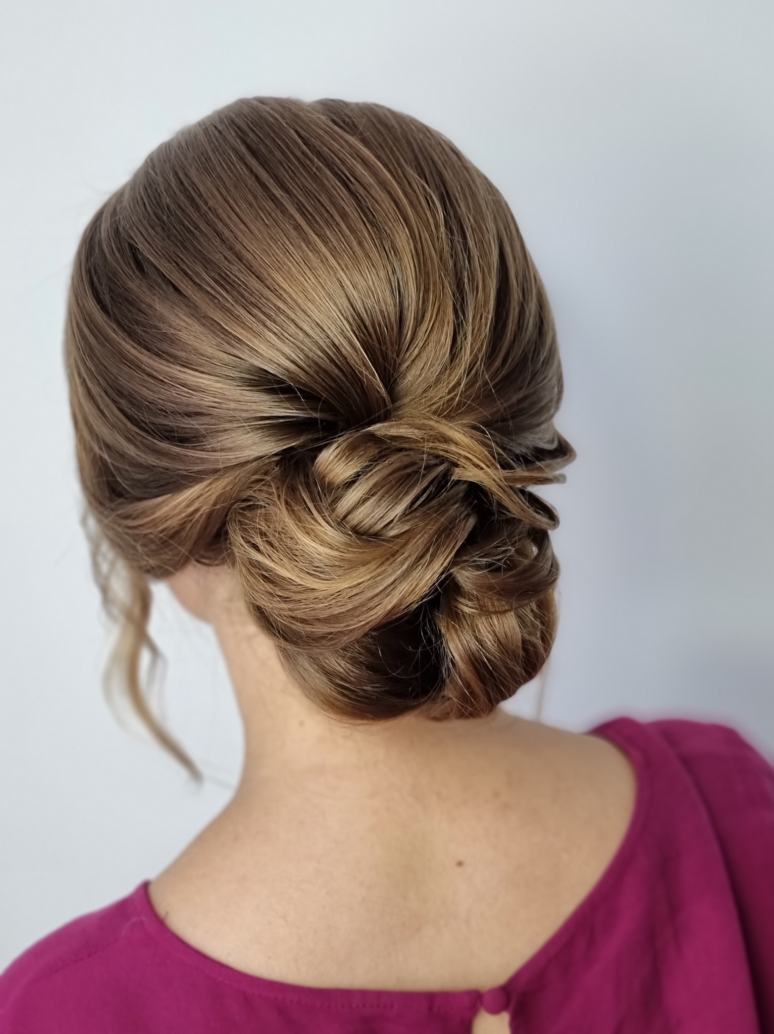 Eleganckie upięcie włosów w odcieniach blondu, widoczne od tyłu, na tle jasnej ściany. Kok z misternie splecionych pasm, podkreślający kobiecą stylizację.
