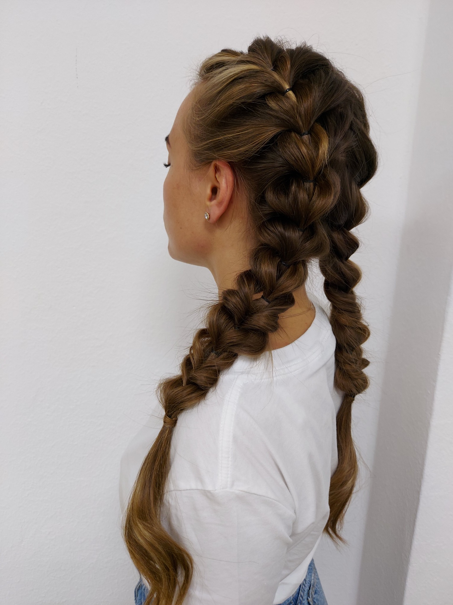 Profil kobiety z upięciem włosów: zaplecione dwa warkocze dobierane, przechodzące w luźne, skręcone pasma. Jasne refleksy na brązowych włosach. Subtelny kolczyk w uchu.