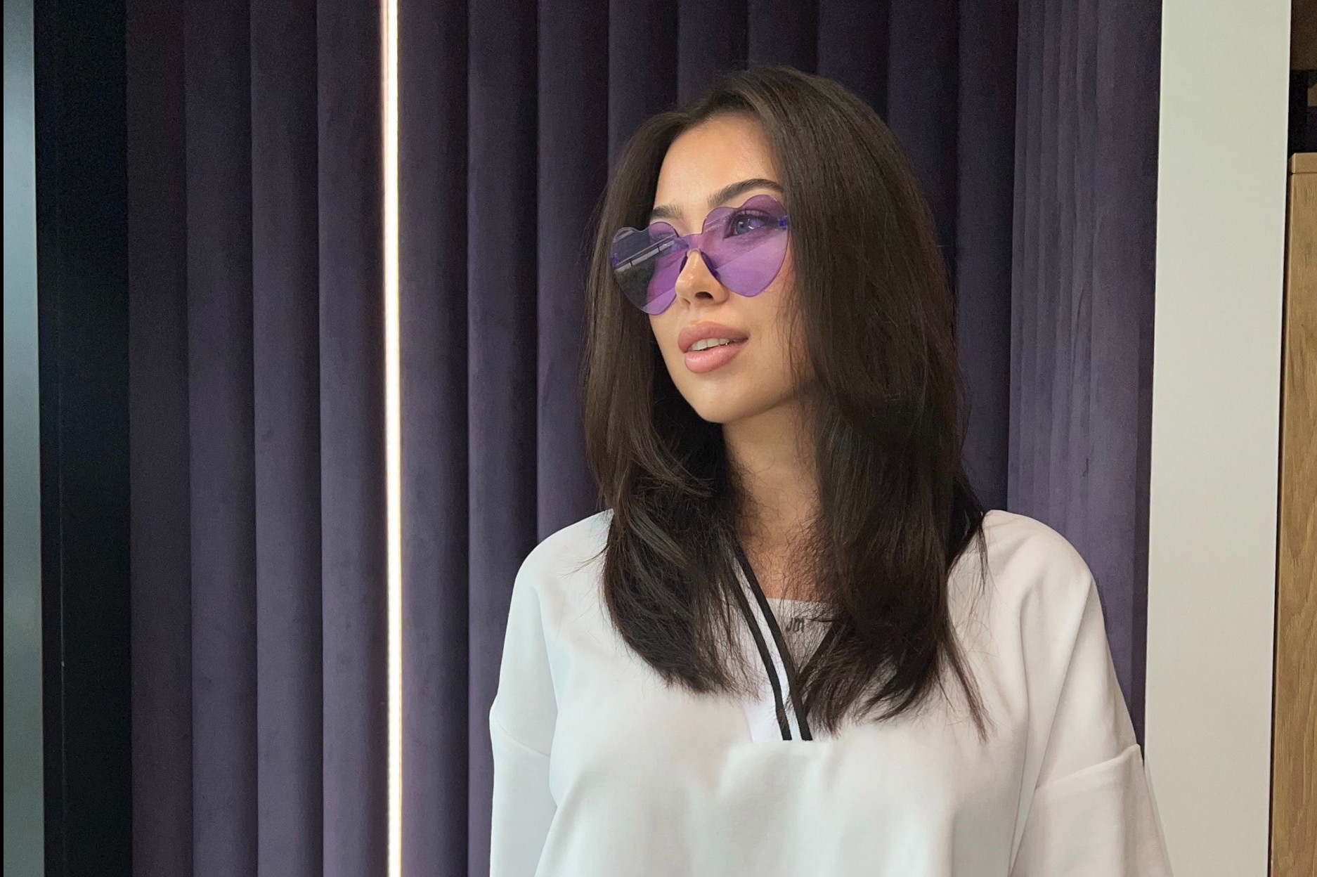 Portret kobiety w okularach w kształcie serc na tle zasłon. Uczesanie z przedziałkiem, delikatny makijaż, jasna bluzka. Ujęcie z boku, oświetlenie naturalne.