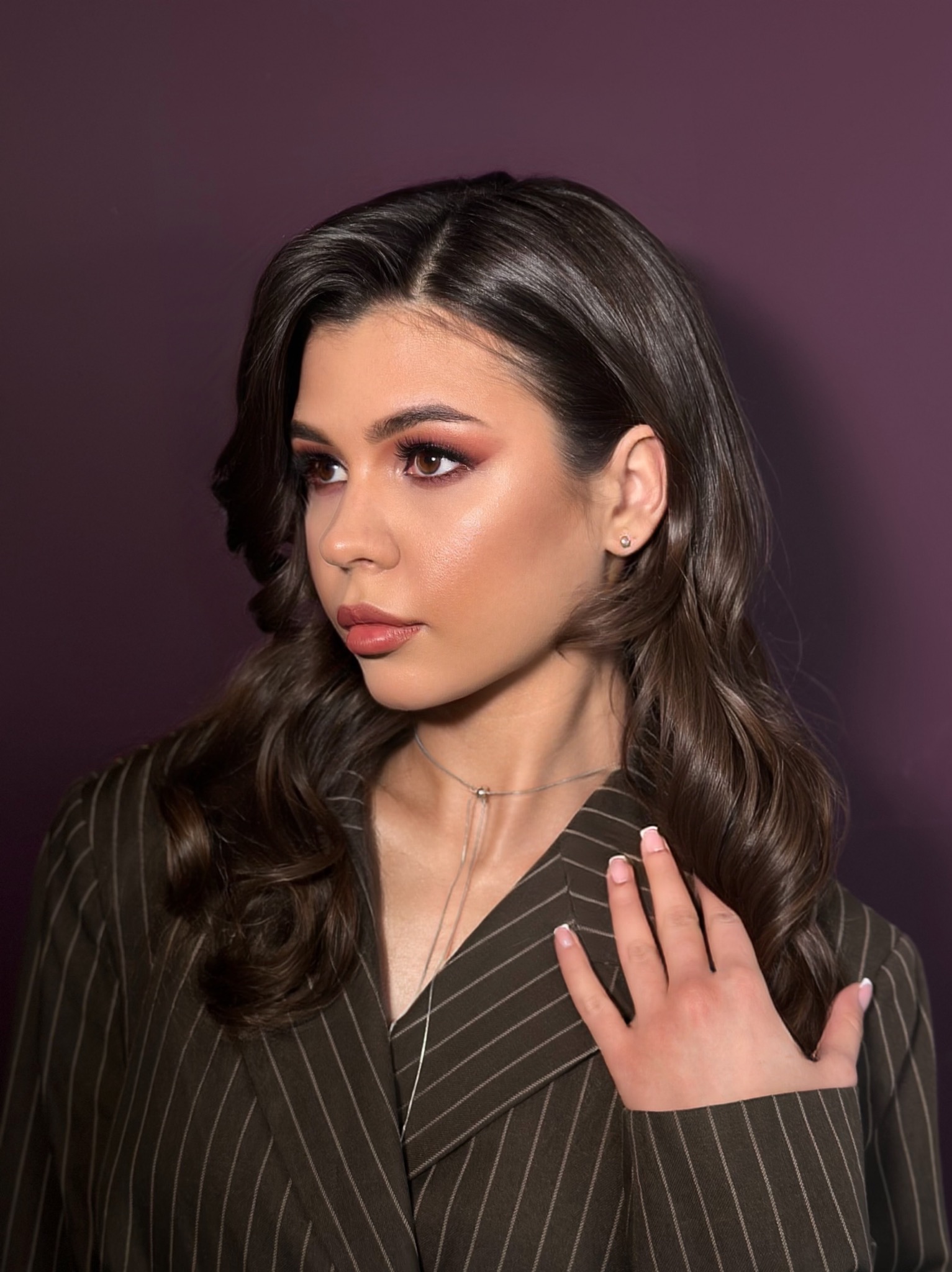 Portret kobiety z profesjonalnym makijażem oczu i ust, brązowe włosy ułożone w fale, ręka na ramieniu w brązowym żakiecie w prążki, fioletowe tło.