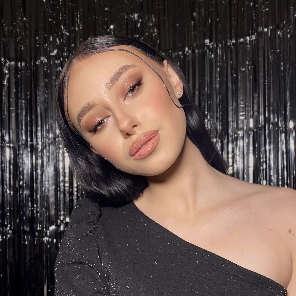 Portret kobiety z delikatnym makijażem w odcieniach nude i błyszczącej, czarnej sukience na tle błyszczącej, czarnej kurtyny.