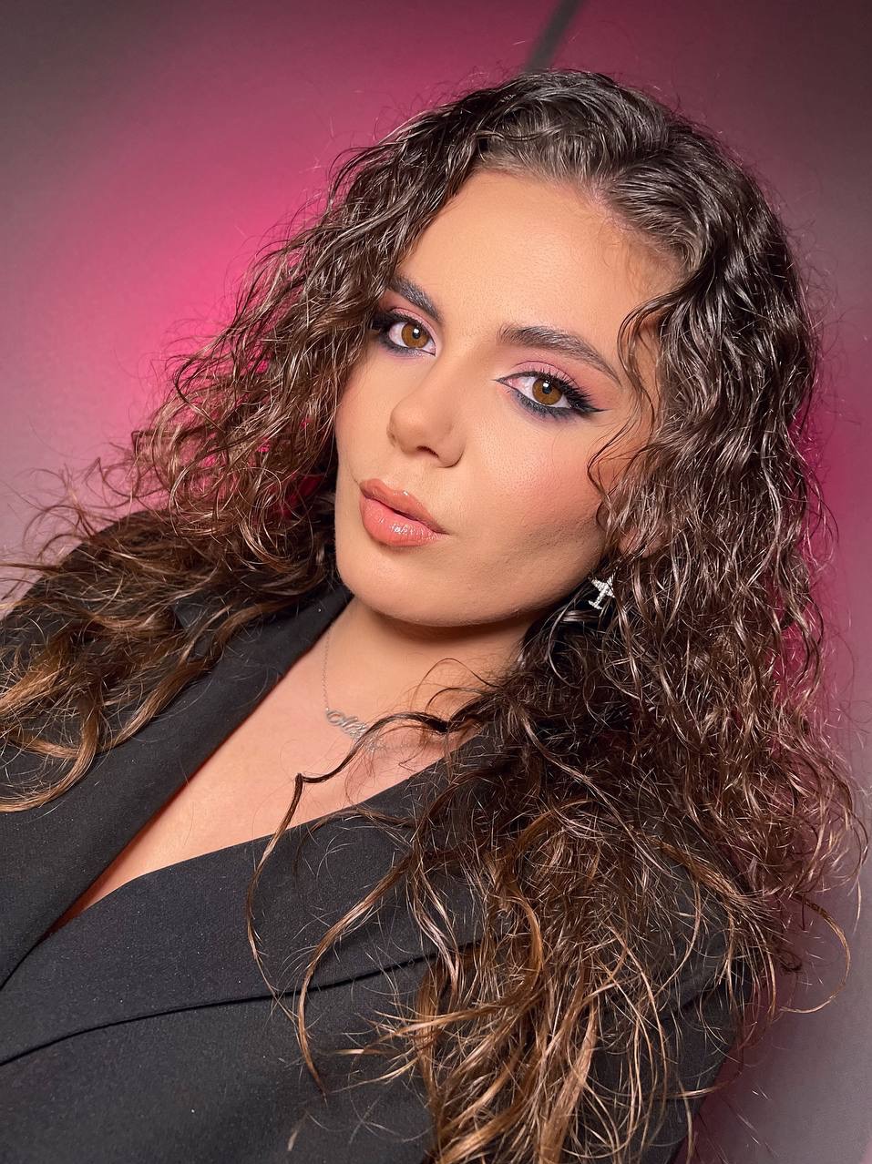 Portret kobiety z burzą kręconych włosów, w czarnej marynarce, z profesjonalnym makijażem oczu i ust, na rozmytym, różowym tle.