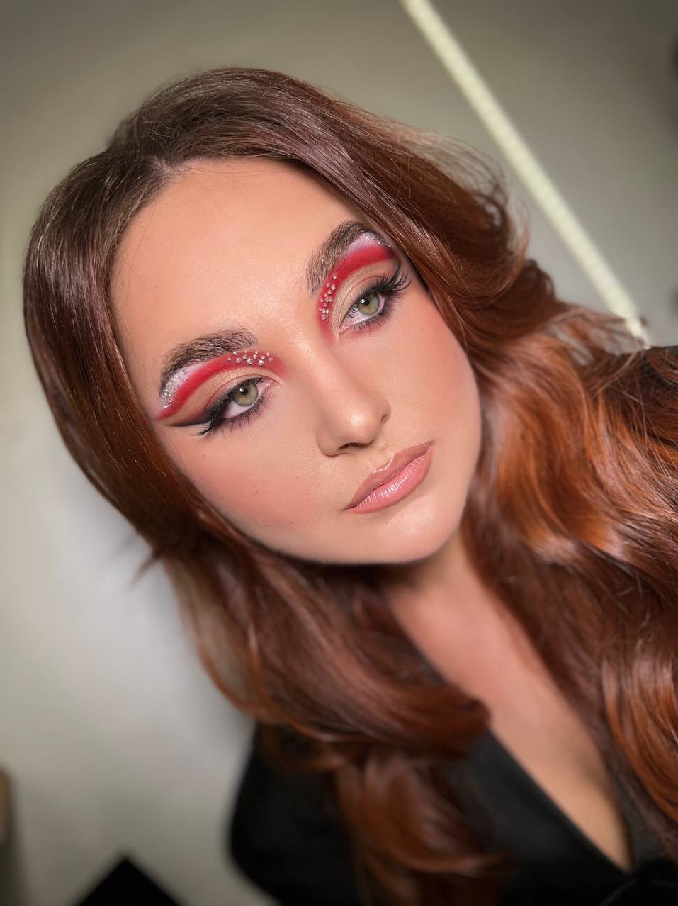 Portret kobiety z artystycznym makijażem oczu w odcieniach czerwieni i srebra, ozdobionym drobnymi kryształkami. Usta błyszczące, włosy długie, rude, pofalowane.