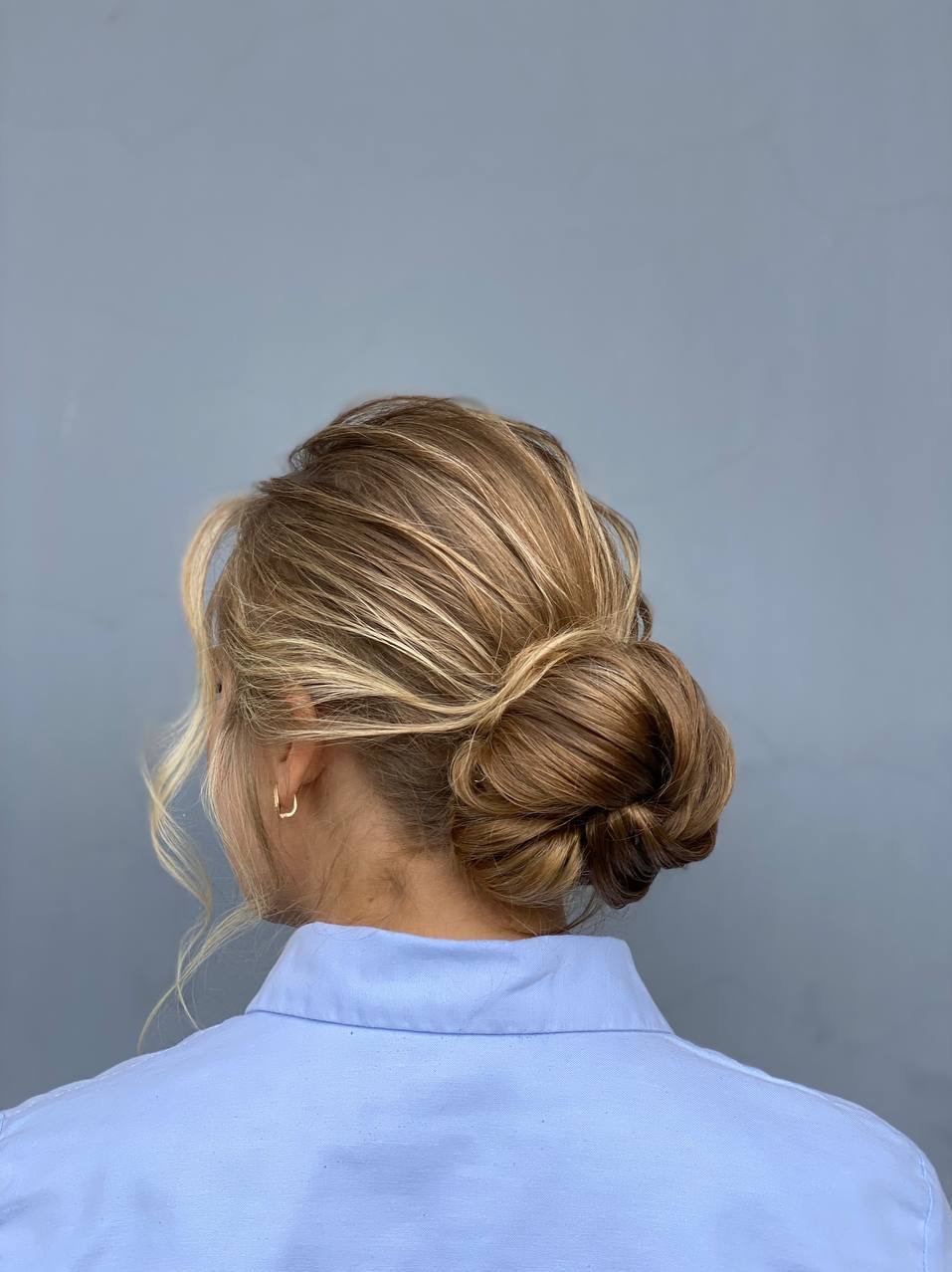 Eleganckie upięcie blond włosów z pasemkami, widok z tyłu. Upięcie typu kok, z delikatnie wypuszczonymi kosmykami. Modelka ma na sobie błękitną koszulę.