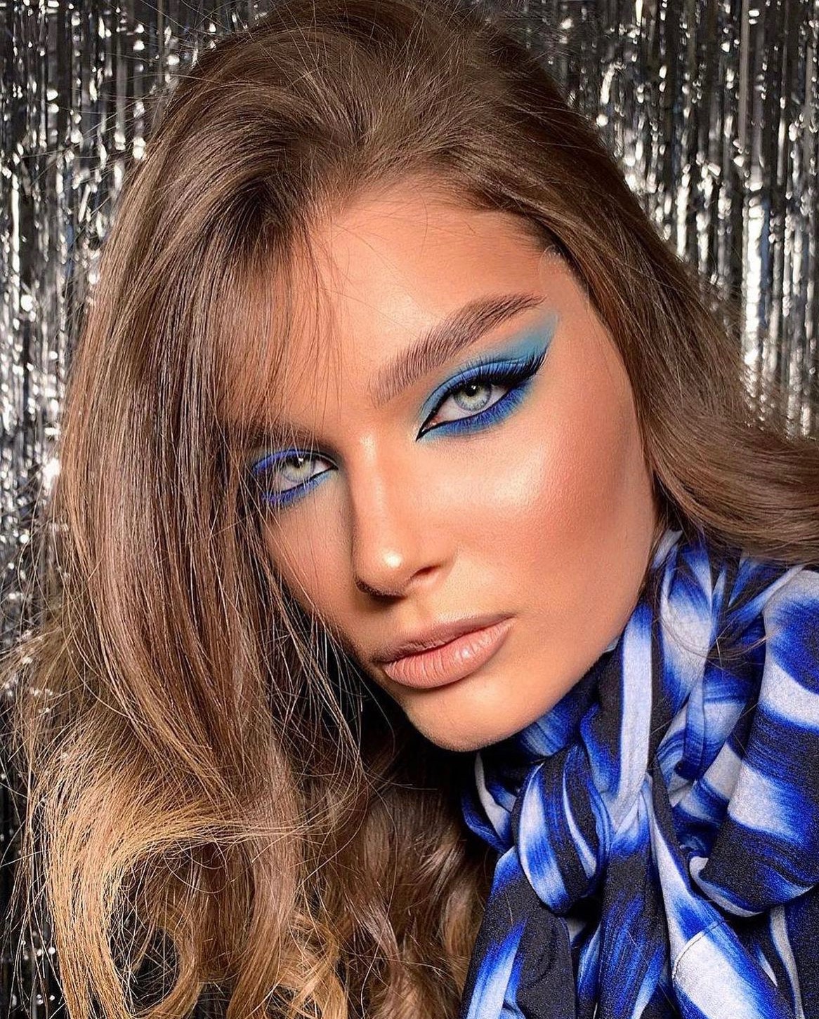 Portret kobiety z intensywnym, niebieskim makijażem oczu i ustami w odcieniu nude, na tle srebrnej, błyszczącej kurtyny. Uwagę zwraca precyzjna kreska i cieniowanie.