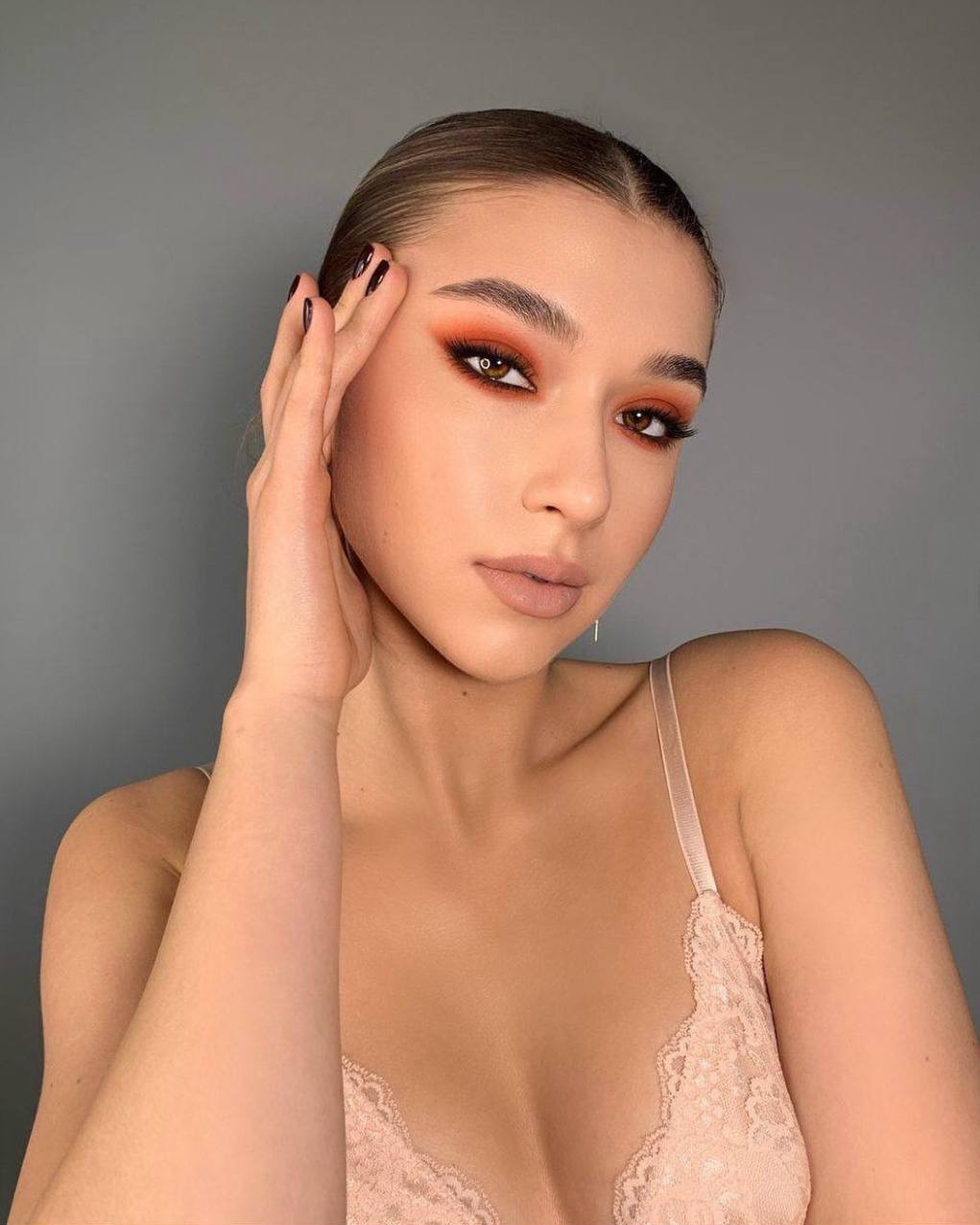 Portret kobiety z intensywnym, pomarańczowym makijażem oczu i subtelnym makijażem ust, pozującej na szarym tle. Widoczne ciemne paznokcie i delikatna biżuteria.