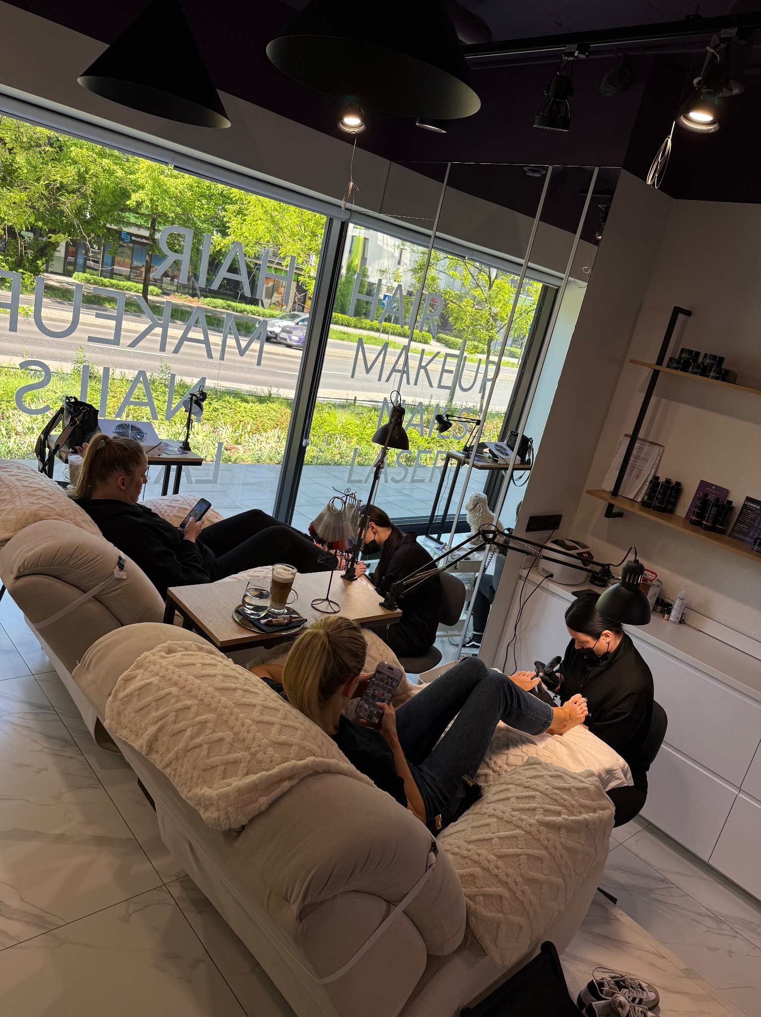 Wnętrze salonu z relaksującą atmosferą. Klientki na sofach, jedna z nich podczas zabiegu pedicure. Duże okno z napisem 'Hair, Nails, Makeup, Laser'.