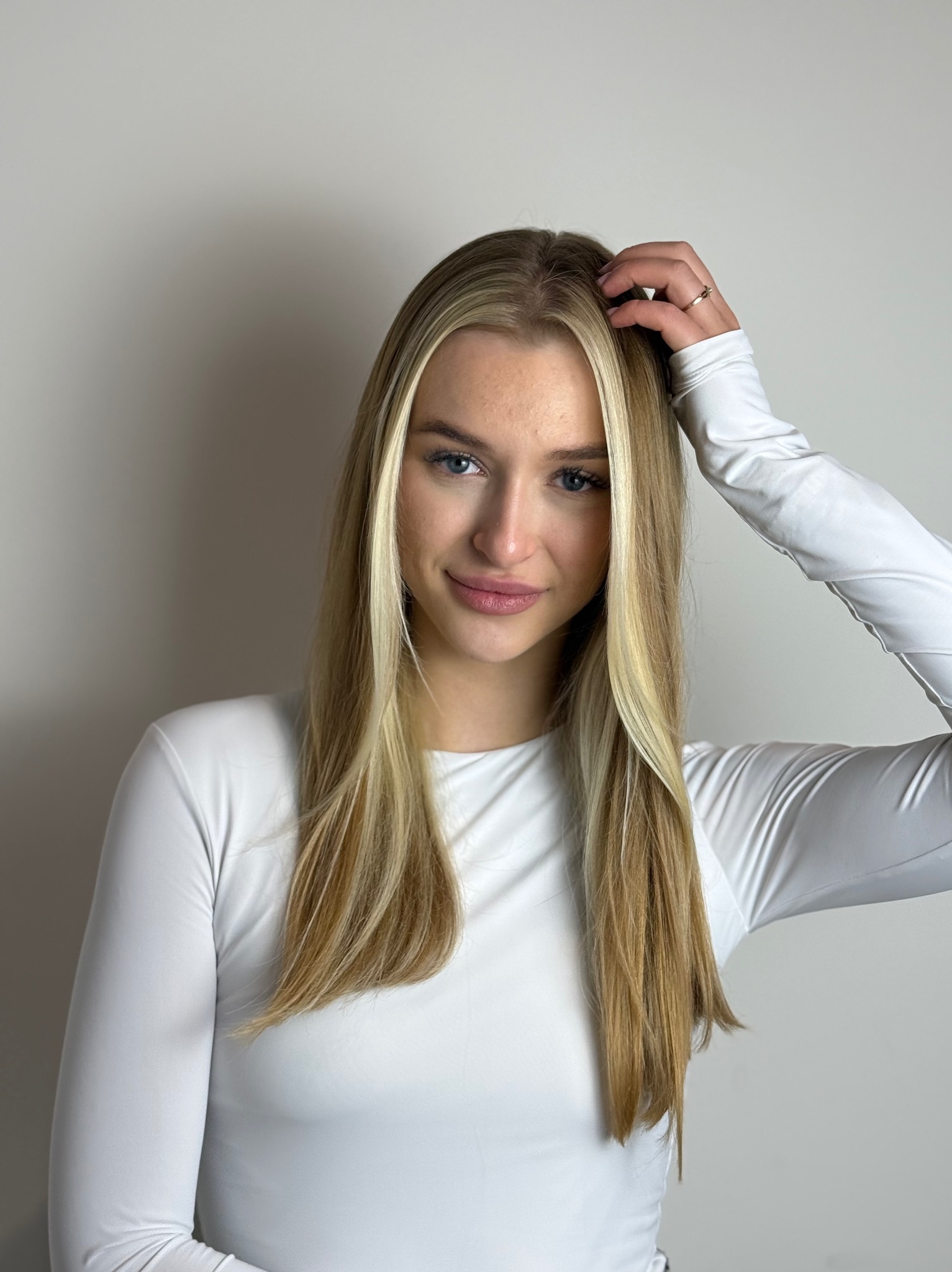 Portret młodej kobiety z blond włosami z pasemkami, w białej bluzce z długim rękawem, dotykającej włosów. Subtelny uśmiech i delikatny makijaż.