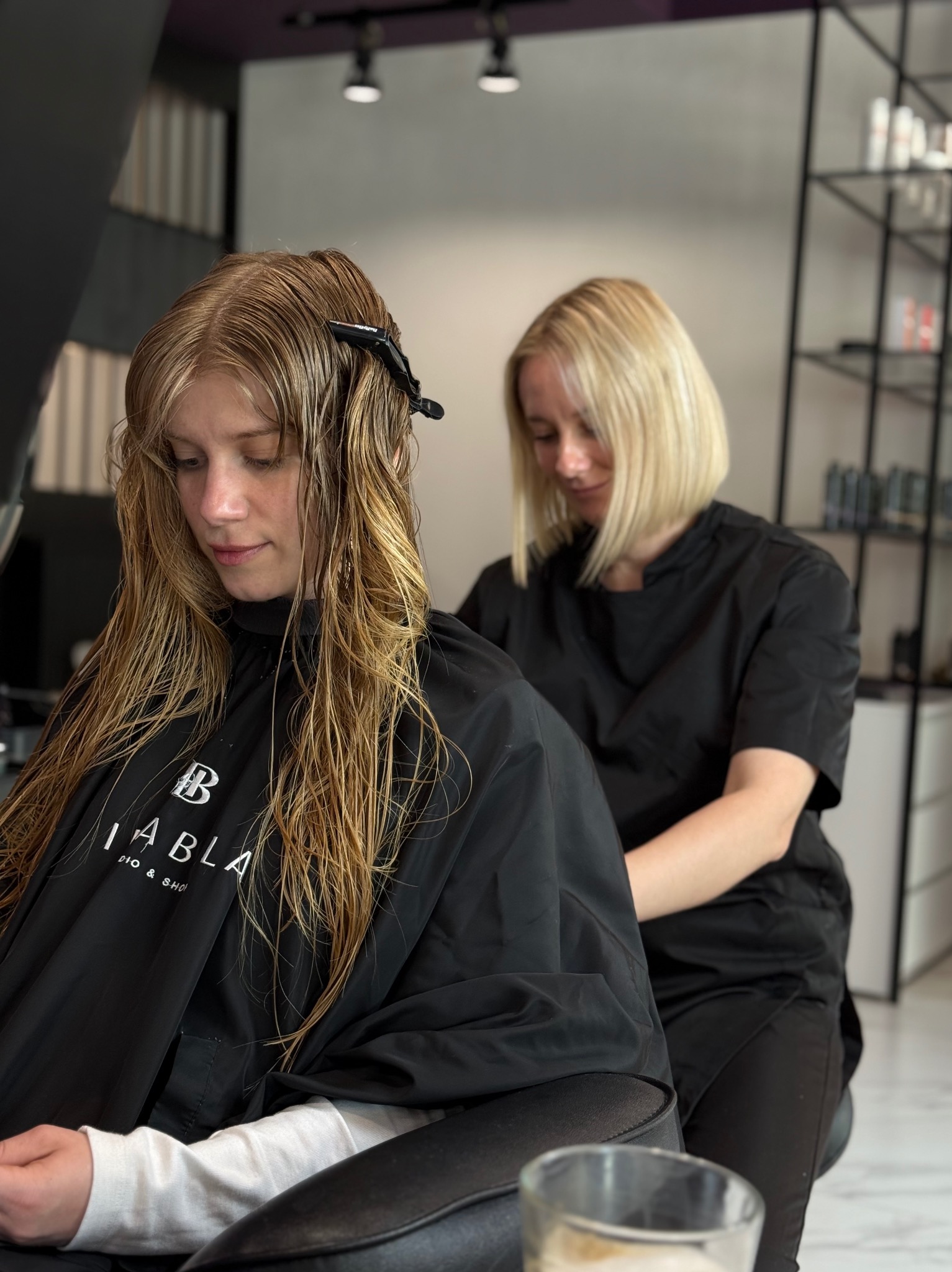 Fryzjerka w czarnym uniformie strzyże mokre, blond włosy klientki w salonie. Na włosach spinka. W tle półki z kosmetykami.