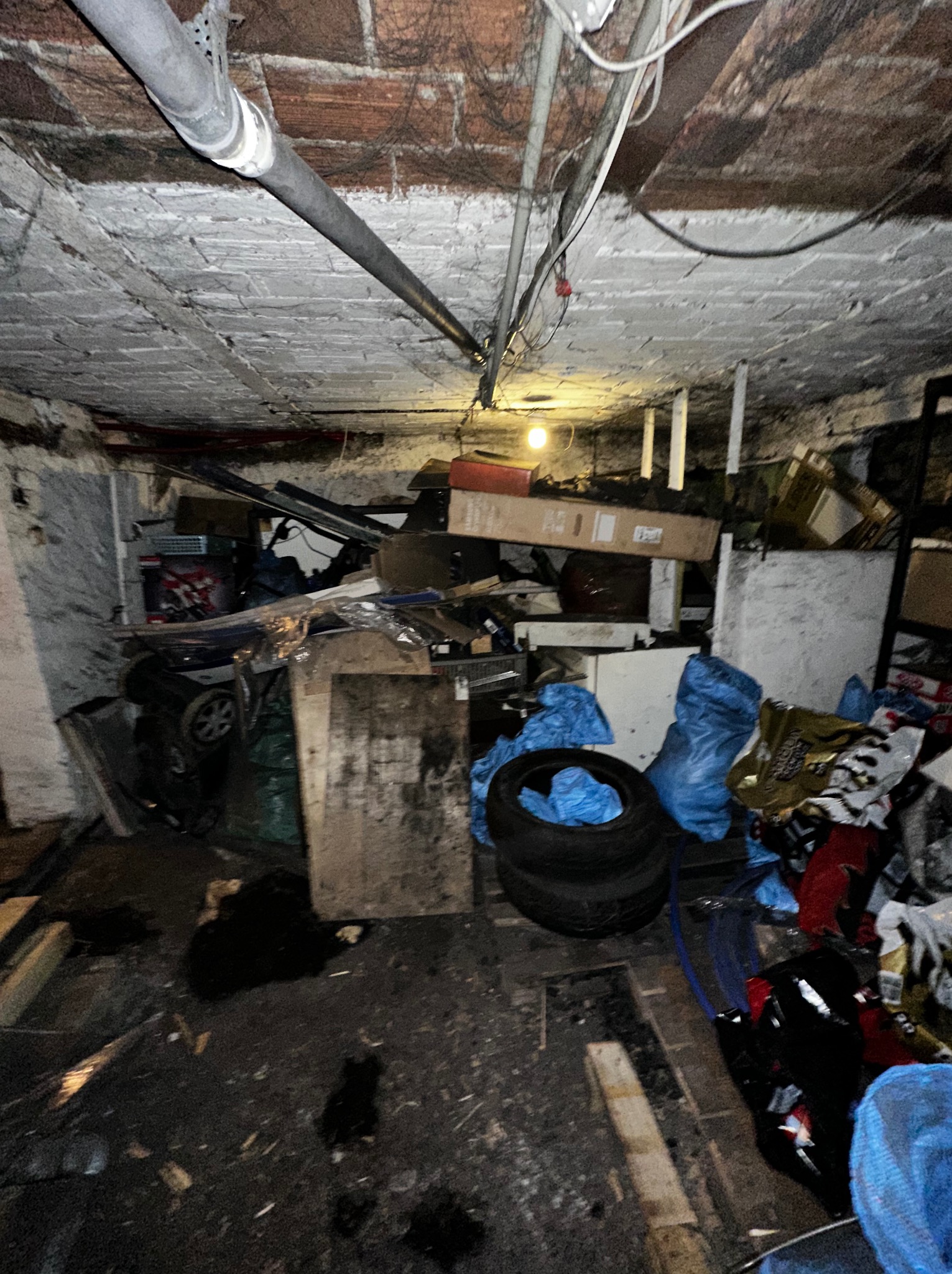 Zagracone, ciemne pomieszczenie z ceglanym sufitem i rurami. Wnętrze wypełnione kartonami, workami i różnymi przedmiotami. Nierówna podłoga z widocznymi deskami.
