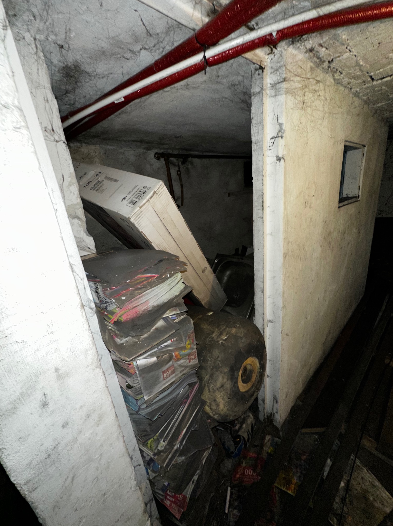 Zagracone pomieszczenie z stertą gazet, kartonem i starą umywalką. Widoczne rury pod sufitem i małe okienko. Niska jakość oświetlenia, brudne ściany.