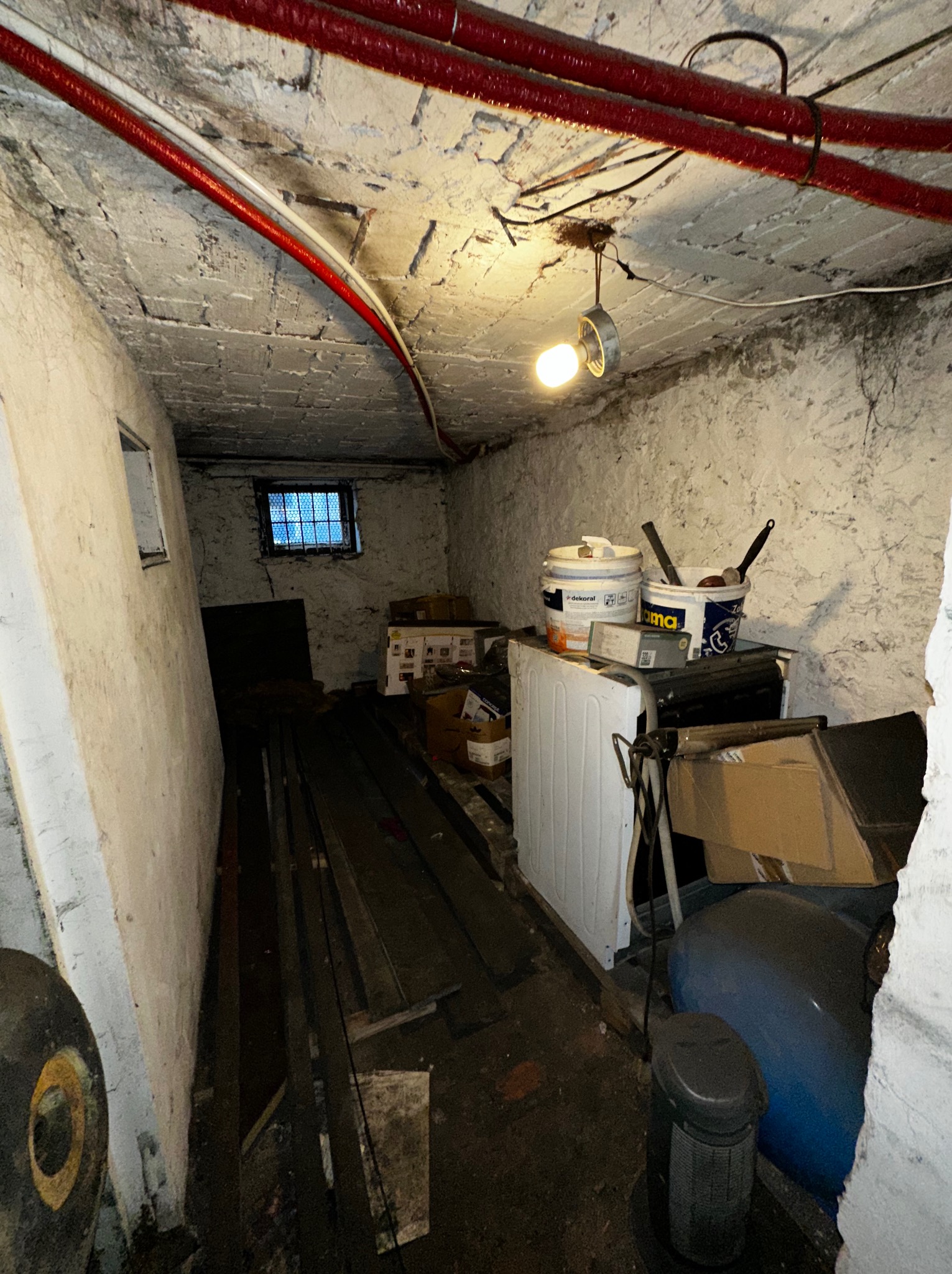 Zagracony, słabo oświetlony piwniczny schowek z wiadrami farby, starą pralką i różnymi przedmiotami. Widoczne rury pod sufitem i małe okienko z kratą.