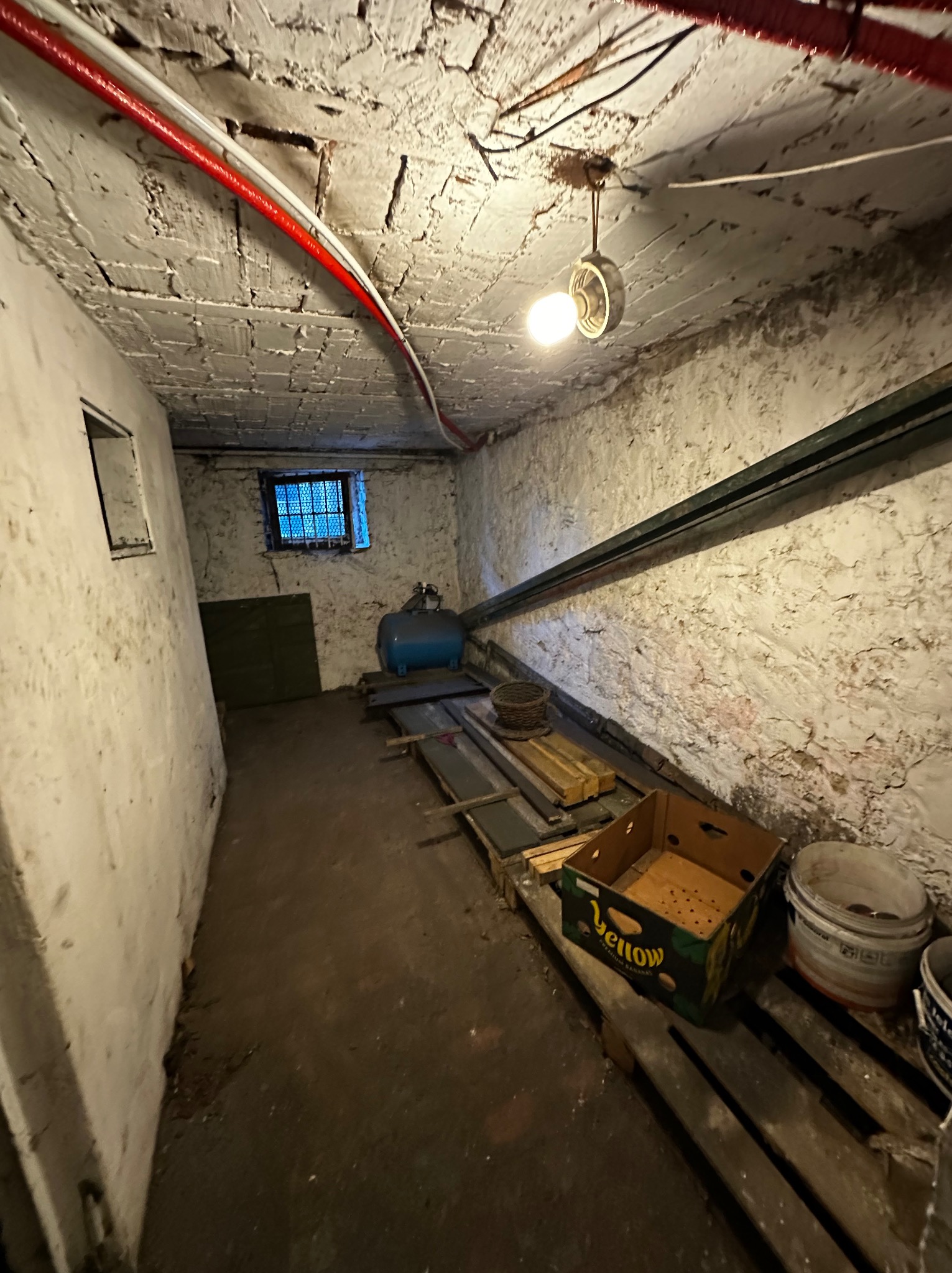 Zaniedbane wnętrze piwnicy z surowymi ścianami, oświetlone pojedynczą żarówką. Widoczne stare rury, skrzynki i inne przedmioty, tworzące wrażenie zagraconej przestrzeni.