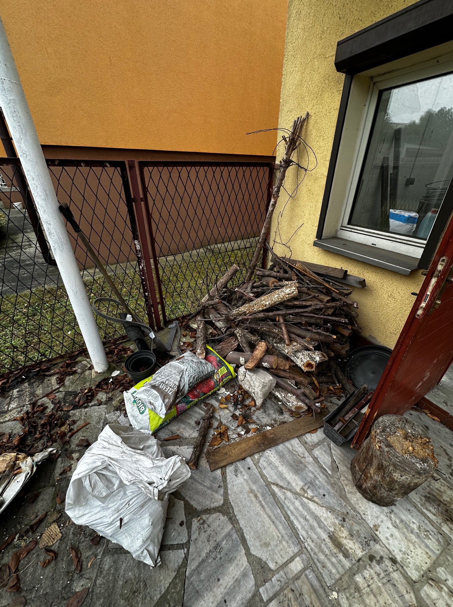 Sterta gałęzi, worki ziemi i narzędzia ogrodnicze przy żółtej ścianie budynku. Widoczne liście i resztki roślinne na kamiennej posadzce.