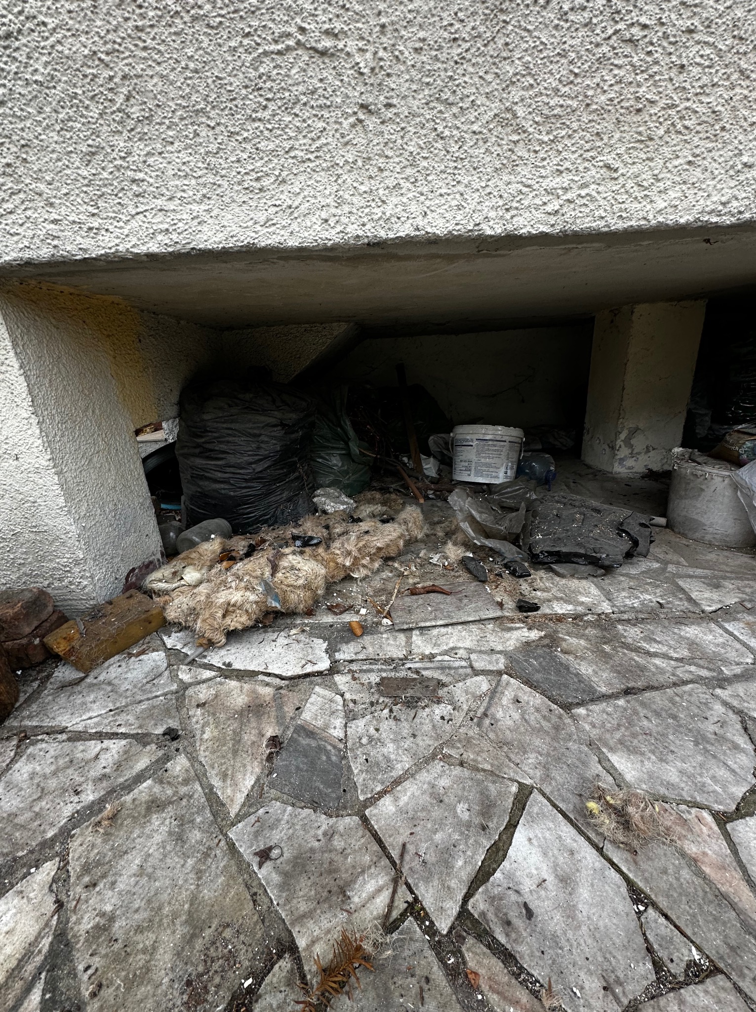Zakamarek z gruzem, workami i odpadami budowlanymi na nierównej, kamiennej posadzce. Widoczne resztki materiałów izolacyjnych i pobrudzone pojemniki.