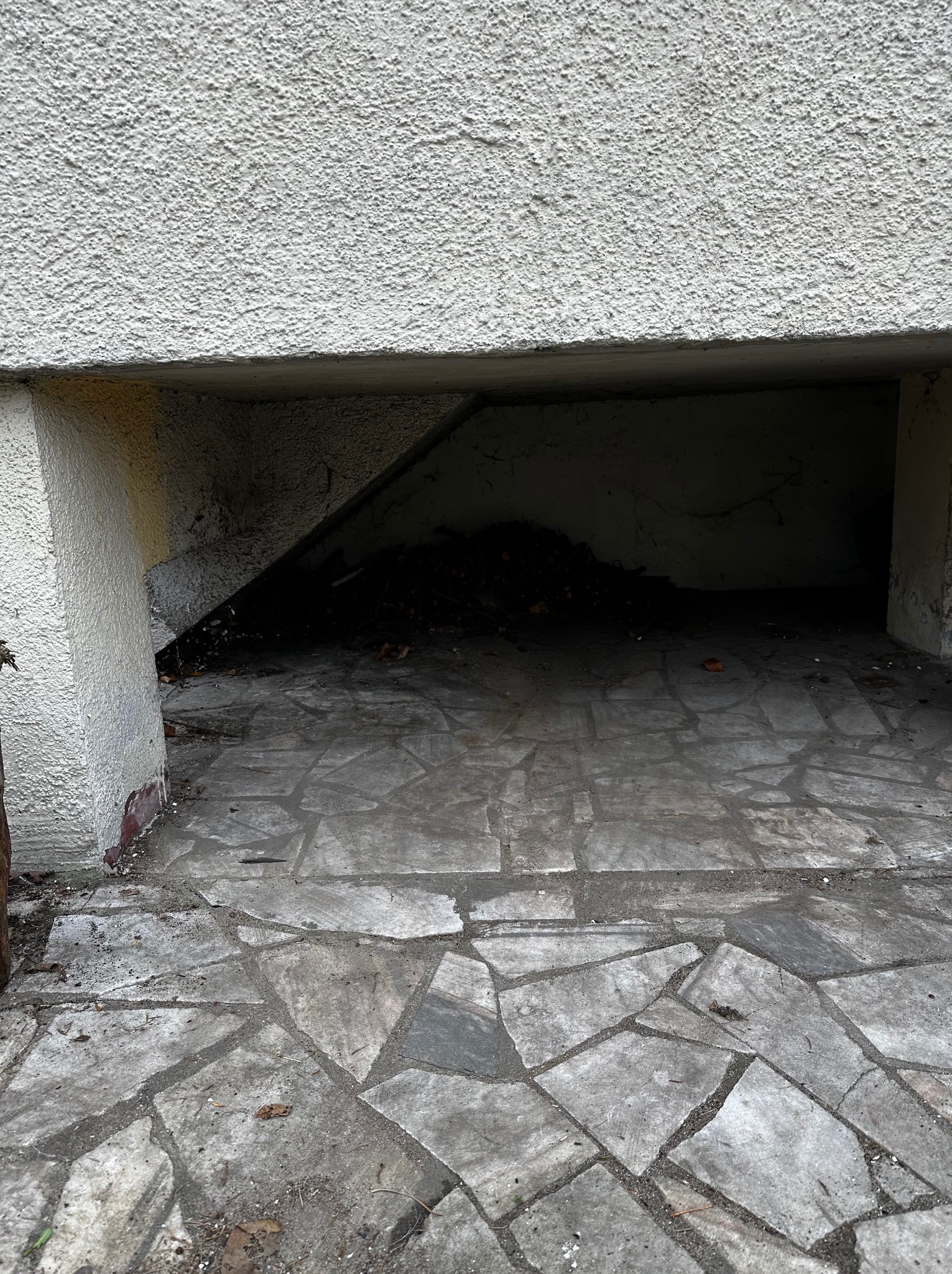 Zakamarek pod schodami z kostką brukową, wypełniony ziemią i liśćmi. Ściany z szorstkiego betonu. Ciemne wnętrze. Nierówna powierzchnia kostki.
