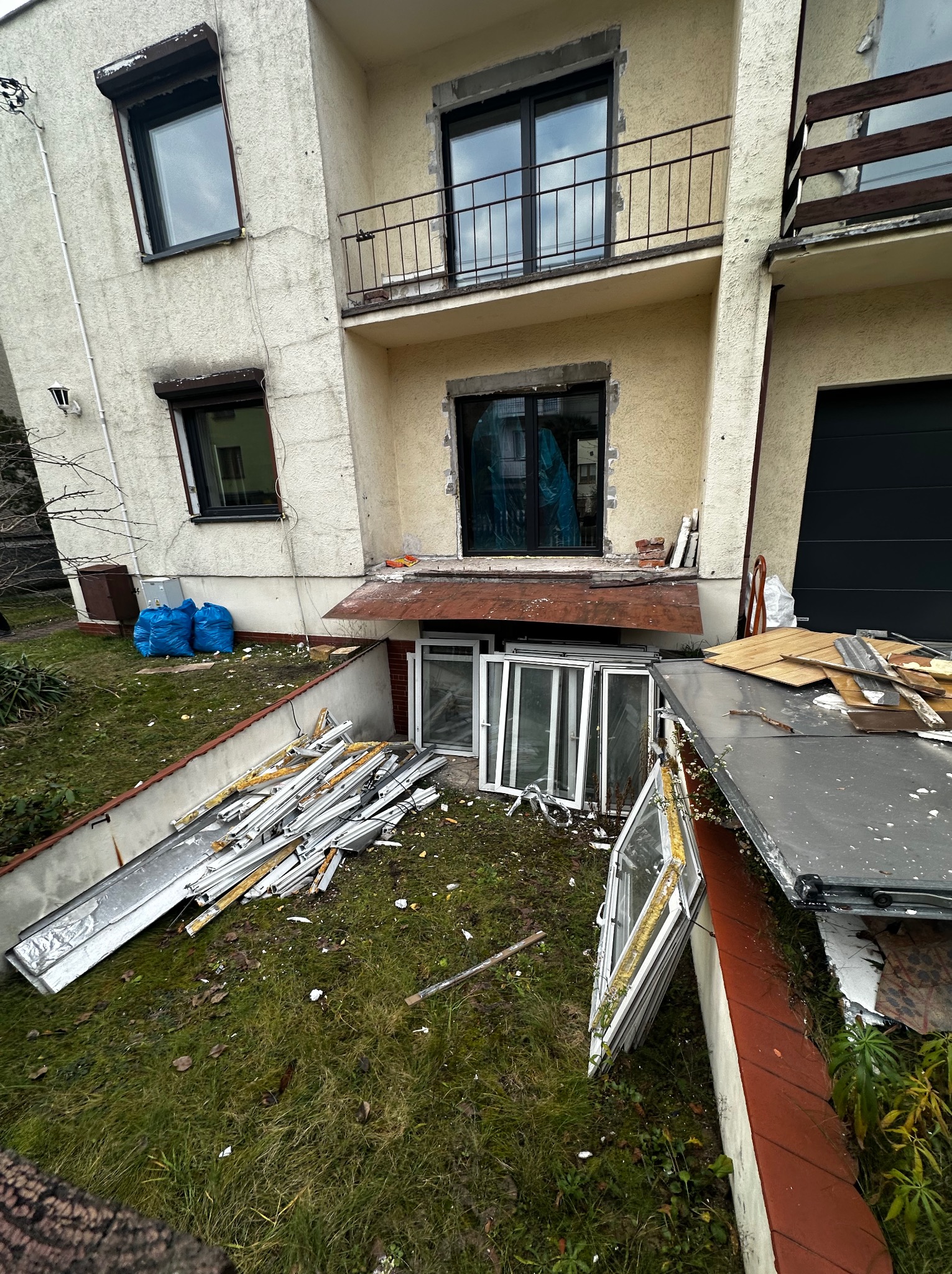 Demontaż okien w budynku. Na trawniku leżą stare ramy okienne i gruz. W oknach widać folię zabezpieczającą. Worki z odpadami przy ścianie budynku.