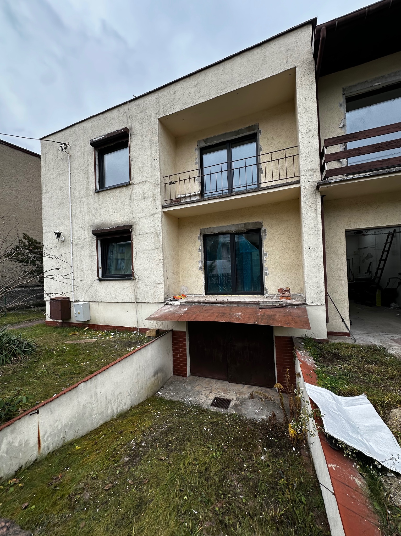 Dwukondygnacyjny dom z balkonami w trakcie remontu elewacji. Widoczne ślady prac, niezabezpieczone okna i bałagan na trawniku. Ujęcie z dołu, pochmurne niebo.