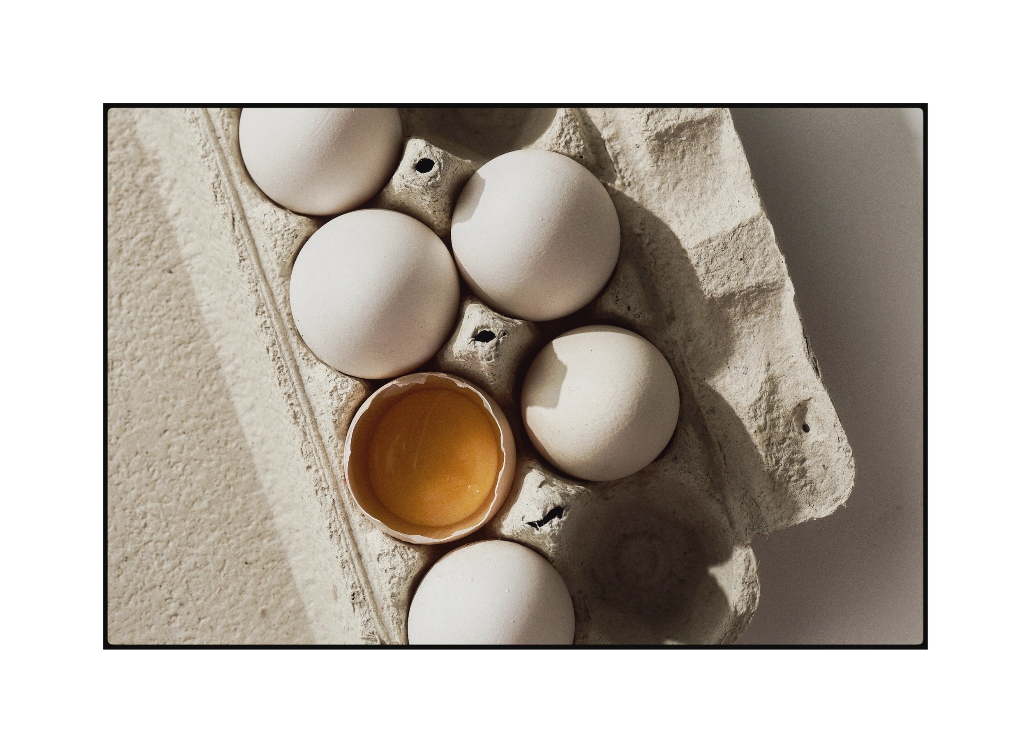 Sześć białych jaj w wytłaczance, jedno rozbite z widocznym żółtkiem, oświetlone naturalnym światłem. Minimalistyczna kompozycja z cieniem.