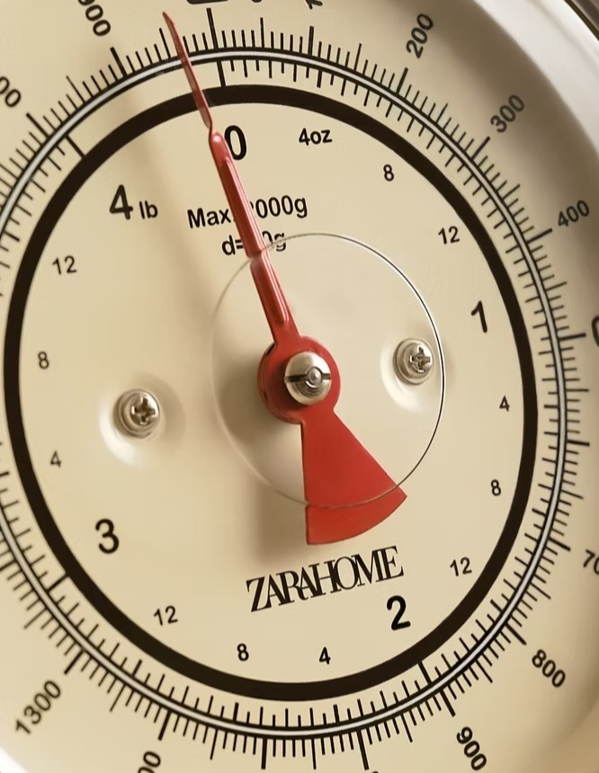 Zbliżenie na tarczę wagi kuchennej w stylu retro z czerwoną wskazówką, skala w kilogramach i funtach, logo Zara Home widoczne na tarczy.