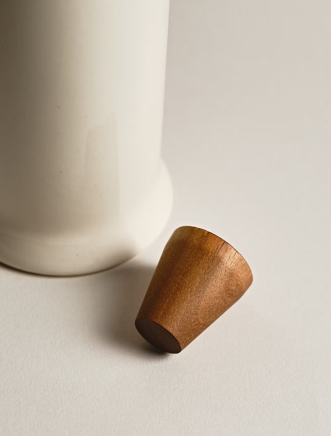 Minimalistyczne ujęcie białego ceramicznego pojemnika z drewnianym korkiem w kształcie stożka na jasnym tle, ukazujące detal i fakturę materiałów.