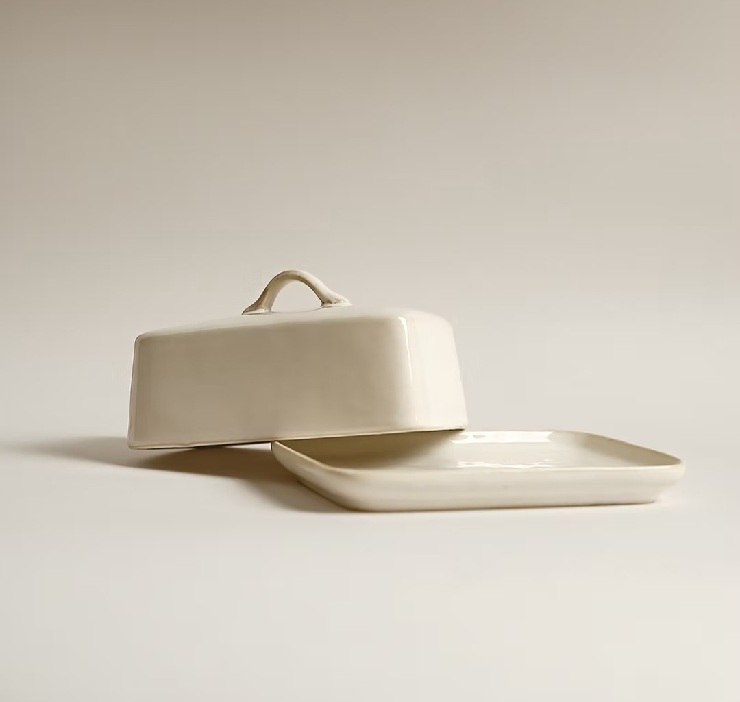 Ceramiczna maselniczka w odcieniach bieli, z pokrywką z uchwytem, ustawiona na talerzyku. Minimalistyczny design i neutralne tło.