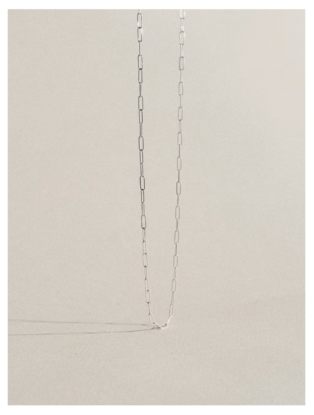 Delikatny srebrny łańcuszek z prostokątnych ogniw na beżowym tle, rzucający subtelny cień. Minimalistyczna biżuteria w neutralnej tonacji.