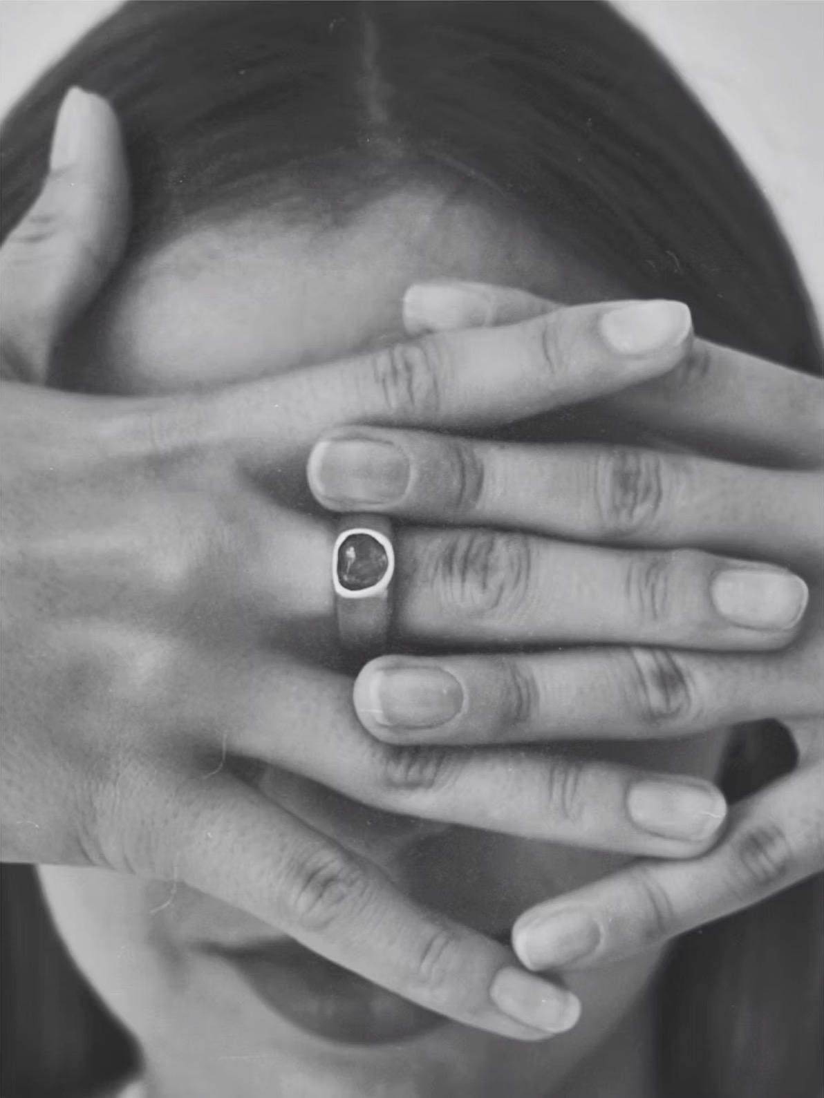 Czarno-białe zbliżenie dłoni kobiety zakrywających twarz, z pierścionkiem z ciemnym kamieniem na palcu. Artystyczne ujęcie, miękkie światło.