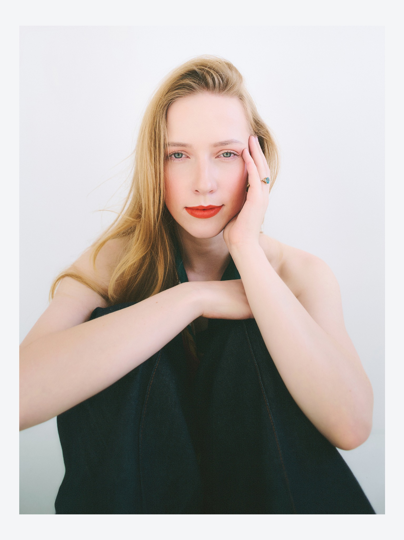 Portret kobiety z blond włosami i czerwonymi ustami, siedzącej na podłodze, z ręką przy twarzy, eksponujący pierścionek z turkusowym oczkiem na tle jasnej ściany.