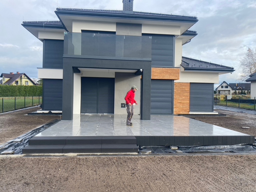 Nowoczesny taras z szarych płyt na podwyższeniu, z widoczną osobą w czerwonej kurtce. Tło stanowi dom z szarymi roletami i elementami drewnianymi.