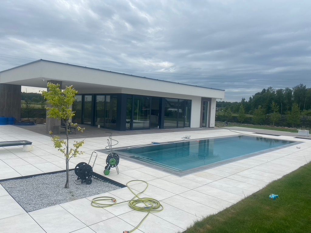 Nowoczesny taras z basenem i jasną kostką brukową. Widoczne narzędzia ogrodowe i młode drzewko. Minimalistyczny styl domu i otoczenia, pochmurne niebo w tle.