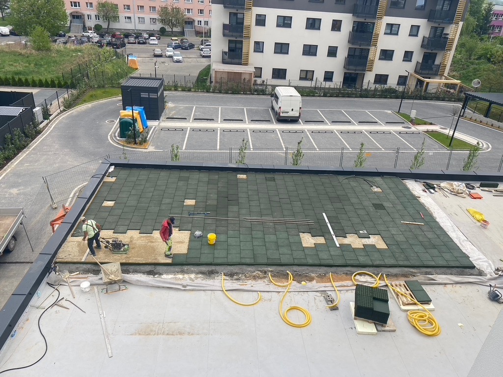 Montaż zielonej nawierzchni z gumowych płyt na dachu budynku; pracownicy używają zagęszczarki. W tle parking i budynki mieszkalne.