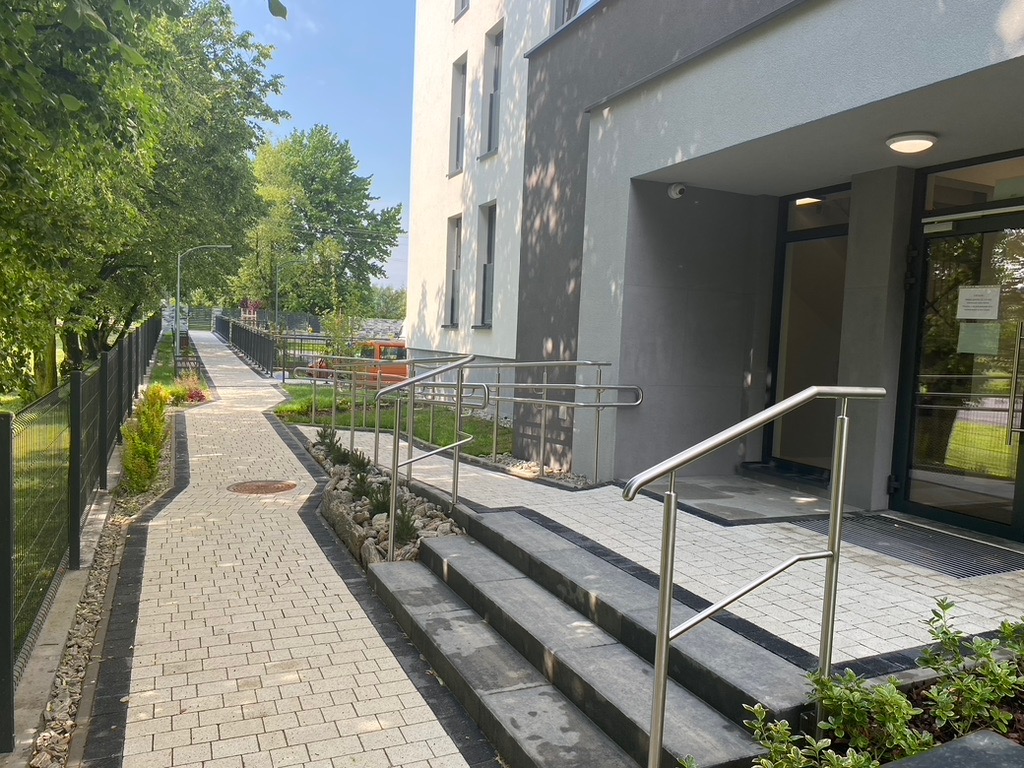 Chodnik z kostki brukowej prowadzący do wejścia budynku, z metalową barierką i ogrodzeniem panelowym wzdłuż ścieżki. Widoczne schody wejściowe i zieleń.