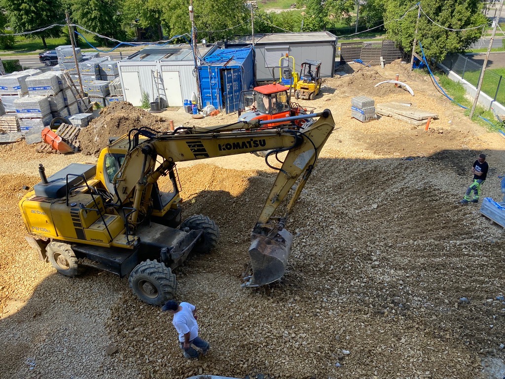 Żółta koparka Komatsu na placu budowy z gruzem, w tle kontenery, palety z kostką i pracownicy. Widok z góry.